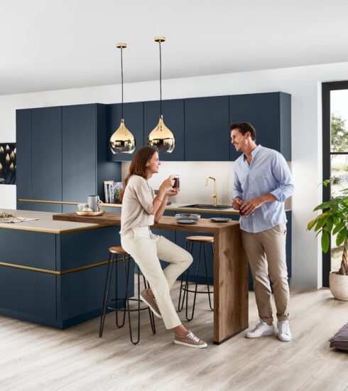 Eine moderne Küchenplanung mit offenen Konzept fördert das kommunikative Zusammenleben. Im Bild ist ein Paar zusehen, dass in einer modernen blauen Küche an der Kücheninsel Frühstücken und sich unterhalten.