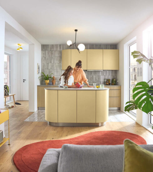 Zwei Frauen an einer modernen, pastellgelben Kücheninsel mit geschwungenen Schränken. Der offene Raum hat einen Holzboden und einen großen, roten Teppich im Vordergrund.