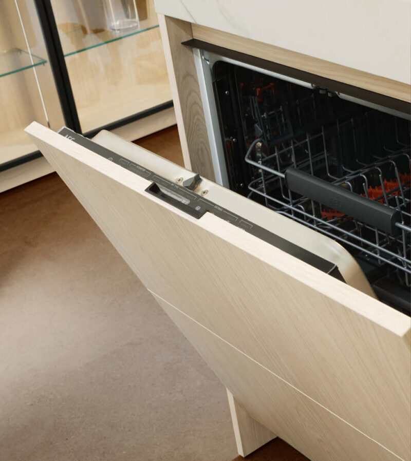 A partially open built-in dishwasher with a light wood-effect panel (likely oak or ash). The control panel is visible at the top edge of the open door, showing the buttons and settings. The stainless steel racks inside the appliance are partially visible.