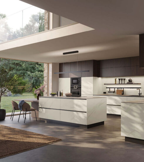 Luxuriöse, moderne Küche mit einer großen Kücheninsel in weißer Marmoroptik. Die Hängeschränke sind dunkelbraun gehalten. Die Küche ist durch eine breite Glasfront zum üppig grünen Garten hin geöffnet.