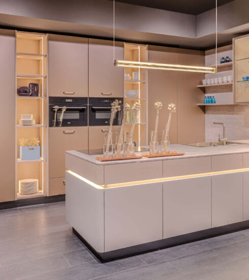 Moderne Küche mit hohen Schränken in warmem Beige/Taupe. Die Kücheninsel und die offenen Regale sind mit indirektem, warmem Licht beleuchtet. Über der Insel hängt eine lange, goldene Pendelleuchte.