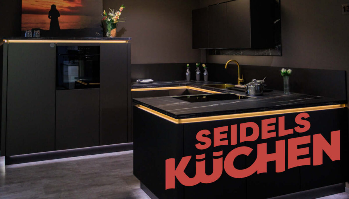 Schwarze U-Küche mit Kehlleistenbeleuchtung. Darauf ist das Logo von Seidels Küchen.