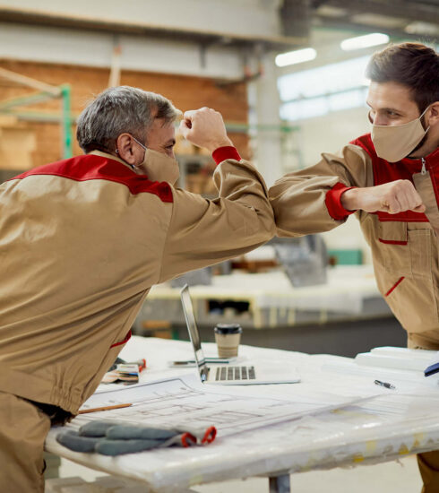 Zwei Handwerker mit Masken im Produktionsbetrieb – gemeinschaftliches Arbeiten in deutscher Küchenfertigung.