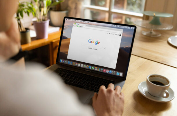 Person sitzt an einem Tisch und nutzt einen Laptop mit geöffneter Google-Startseite; daneben steht eine Tasse Kaffee in heller, wohnlicher Umgebung.