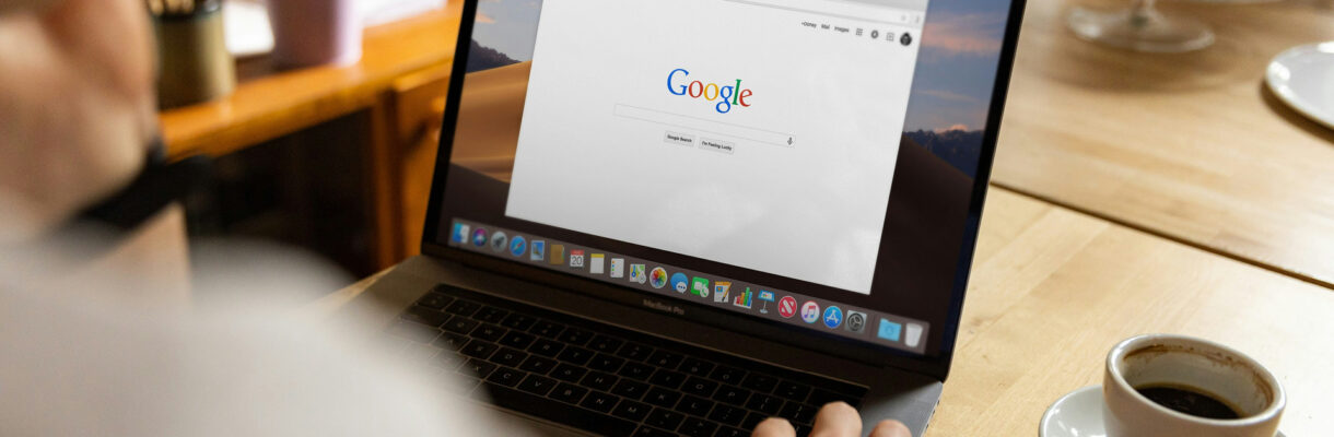 Person sitzt an einem Tisch und nutzt einen Laptop mit geöffneter Google-Startseite; daneben steht eine Tasse Kaffee in heller, wohnlicher Umgebung.