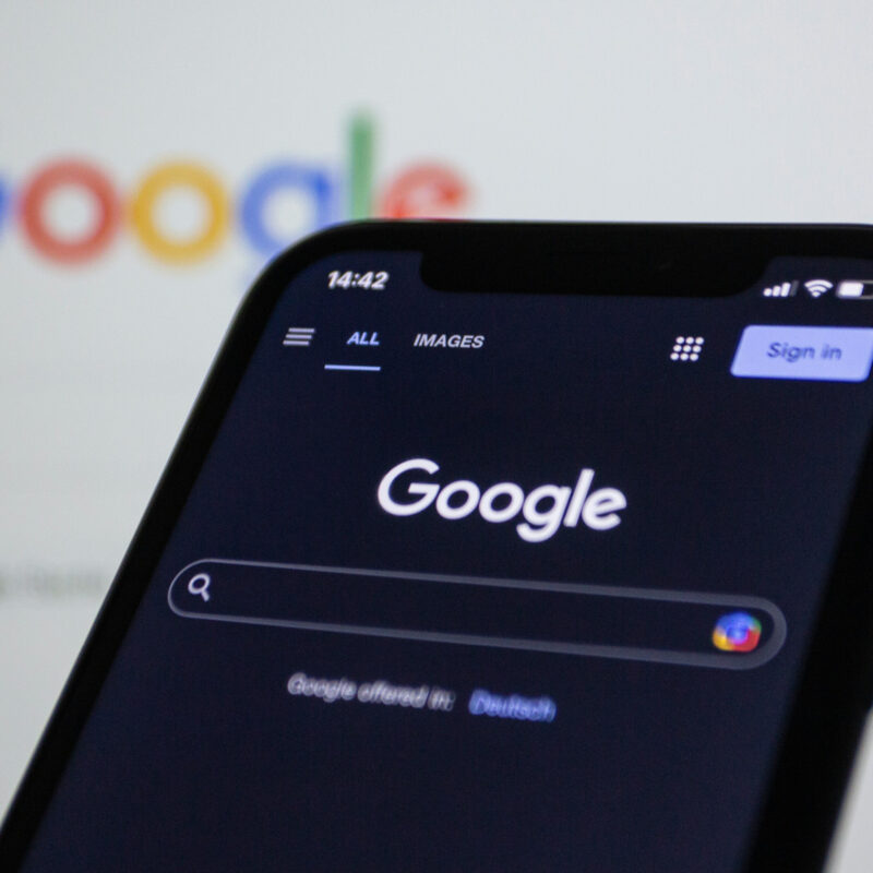 Smartphone mit geöffneter Google-Suchseite im Dunkelmodus, im Hintergrund unscharf das Google-Logo auf einem Bildschirm.