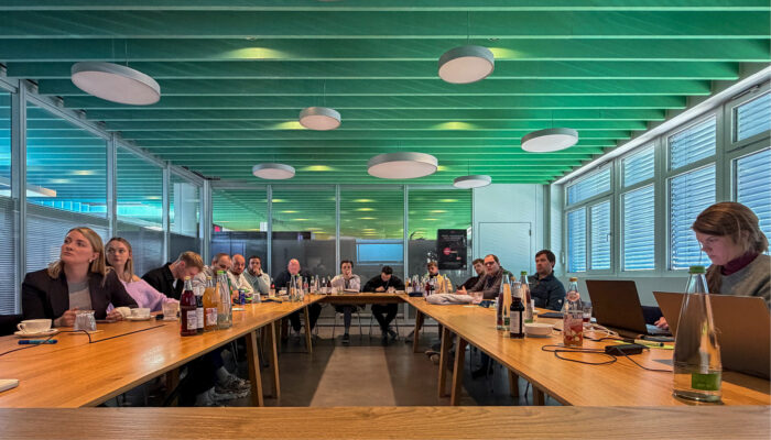 Mehrere Teilnehmende sitzen an einem U-förmig angeordneten Tisch in einem modernen Konferenzraum und verfolgen eine Besprechung.