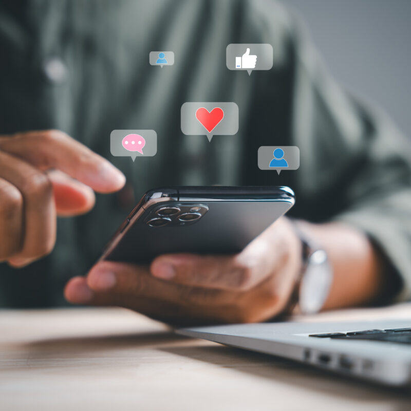 Person nutzt ein Smartphone auf einem Schreibtisch neben einem Laptop. Über dem Gerät schweben Symbole für Likes, Kommentare, Reaktionen und Nutzerinteraktionen, die soziale Netzwerke und digitale Kommunikation darstellen. Das Bild symbolisiert Social-Media-Aktivitäten, Community-Management und den Austausch mit Kunden über digitale Kanäle.