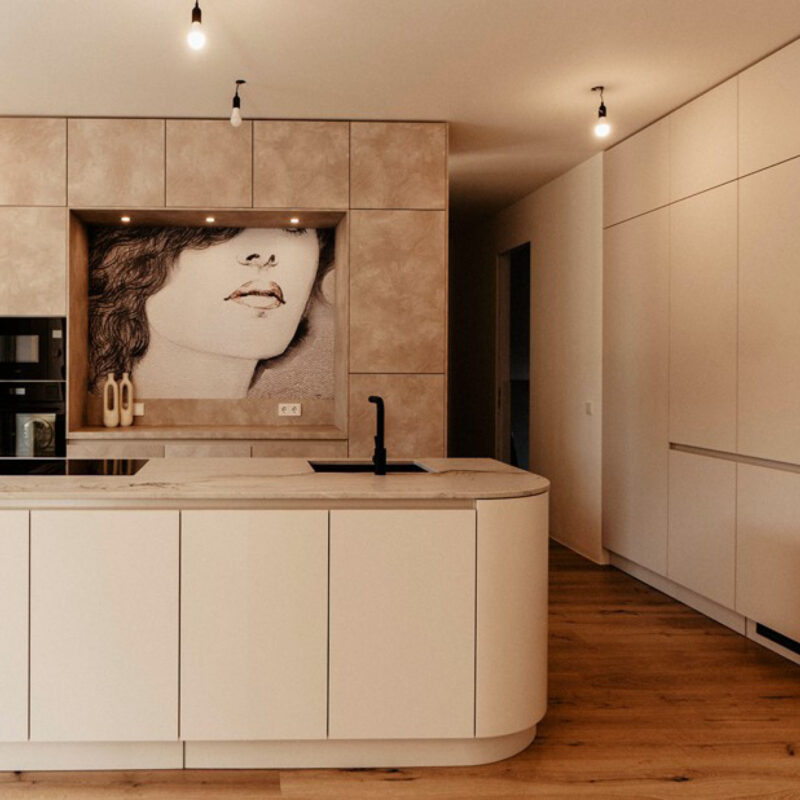 Moderne, grifflose Wohnküche in warmen Beige- und Naturtönen mit Kochinsel, integrierter Spüle und deckenhohen Schränken. Ein großformatiges Kunstmotiv an der Rückwand setzt einen dekorativen Akzent und verleiht der minimalistischen Raumgestaltung eine persönliche Note.
