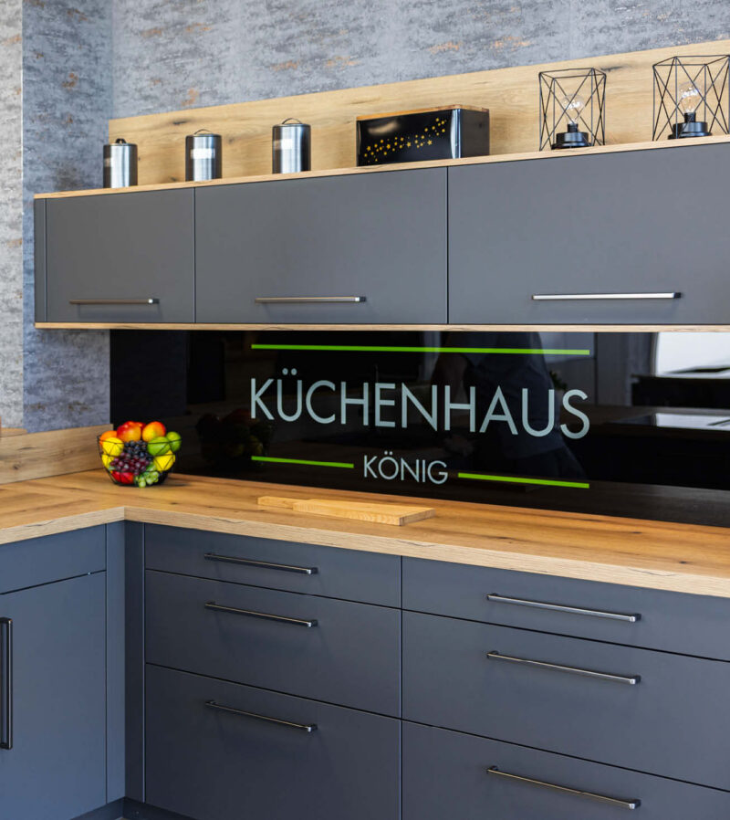 Moderne Ausstellungsküche im Küchenhaus König mit anthrazitfarbenen Küchenfronten, Arbeitsplatte in Holzoptik und hochwertiger Glasrückwand mit Logo. Die Einbauküche verbindet zeitloses Küchendesign, großzügigen Stauraum und individuelle Küchenplanung für moderne Wohnküchen.