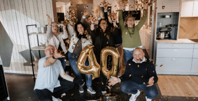Eine Gruppe von neun Personen feiert ein 40-jähriges Jubiläum in einer modernen Küchenausstellung. Goldenes Konfetti fliegt durch die Luft, in der Mitte stehen goldene Luftballons mit der Zahl „40“, während die Menschen lachend die Arme in die Höhe strecken.