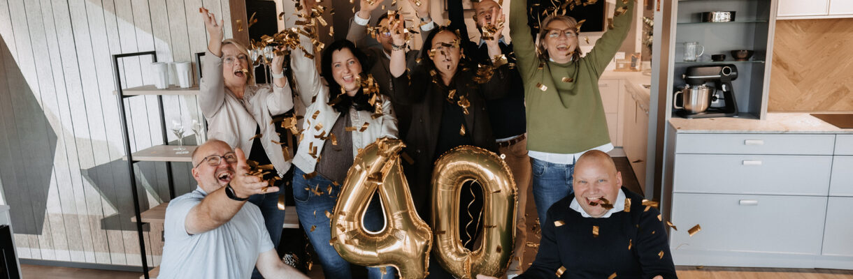 Eine Gruppe von neun Personen feiert ein 40-jähriges Jubiläum in einer modernen Küchenausstellung. Goldenes Konfetti fliegt durch die Luft, in der Mitte stehen goldene Luftballons mit der Zahl „40“, während die Menschen lachend die Arme in die Höhe strecken.