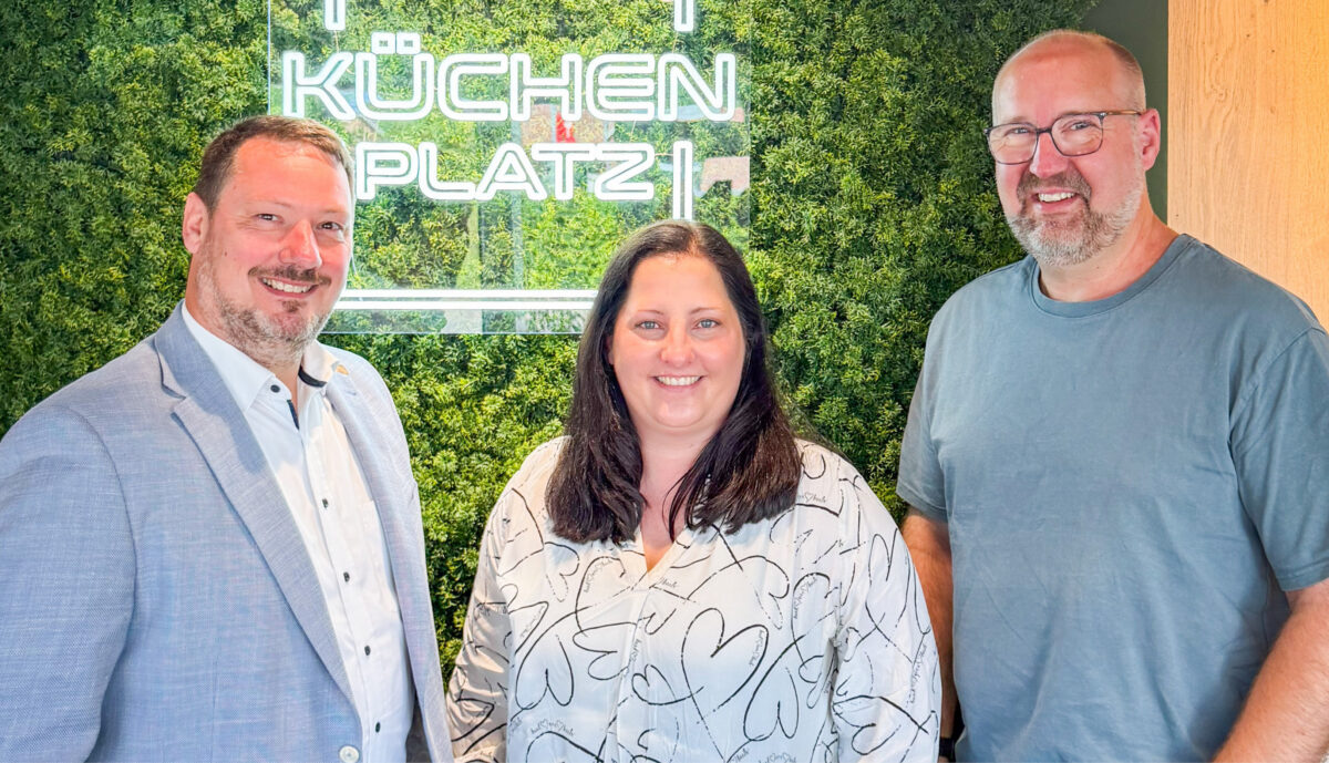 Die Inhaber von Küchen Platz posieren gemeinsam vor dem Firmenlogo im Küchenstudio und stehen für persönliche Beratung, Qualität und langjährige Erfahrung.