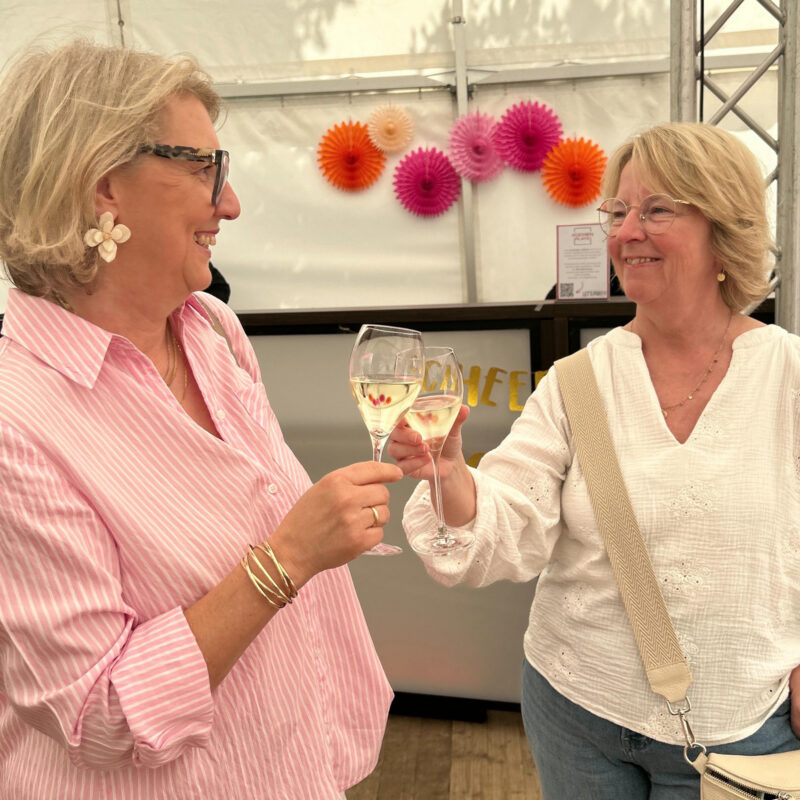 Zwei Frauen stoßen mit Weißweingläsern auf einer Jubiläumsfeier in einem festlich geschmückten Festzelt an und lächeln sich dabei an.