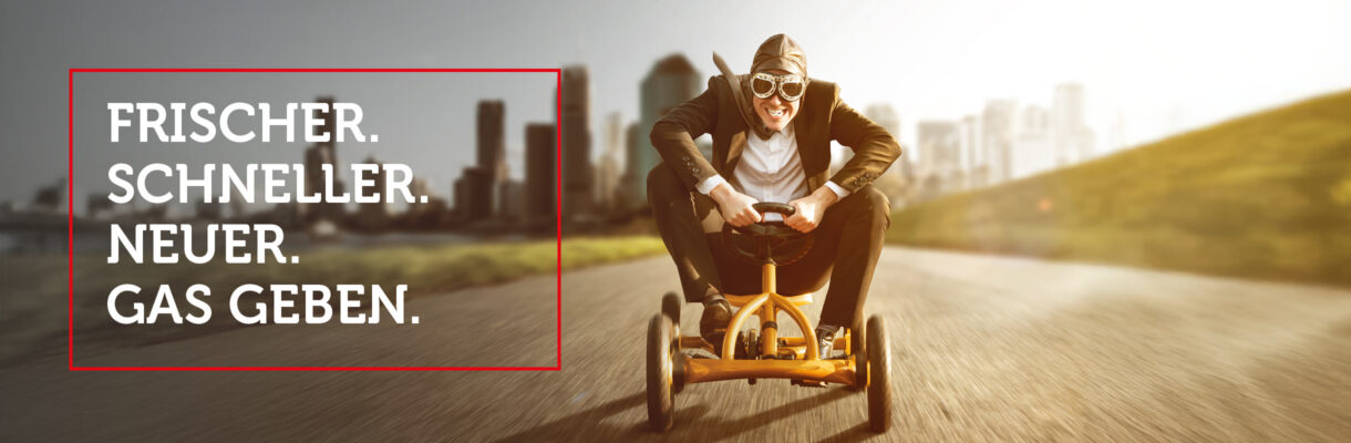 Dynamischer Geschäftsmann mit Fliegerbrille fährt auf einem gelben Pedal-Gokart über eine Straße vor moderner Stadtkulisse – symbolisches Headerbild für innovatives Marketing, Social Media und digitale Sichtbarkeit von Küchenstudios, Küchenplanung und Möbelhandel.