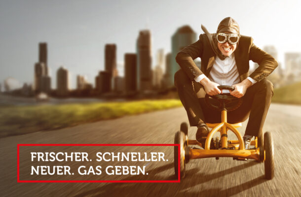 Dynamischer Geschäftsmann mit Fliegerbrille fährt auf einem gelben Pedal-Gokart über eine Straße vor moderner Stadtkulisse – symbolisches Headerbild für innovatives Marketing, Social Media und digitale Sichtbarkeit von Küchenstudios, Küchenplanung und Möbelhandel.