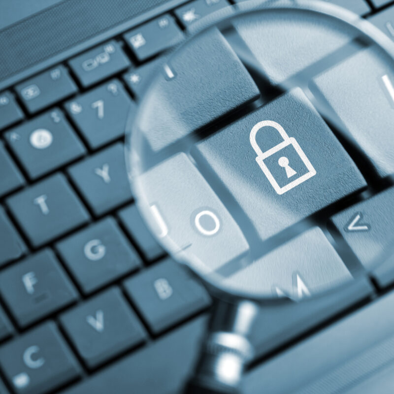 Nahaufnahme einer Computertastatur mit einem Schlosssymbol auf einer Taste, das durch eine Lupe vergrößert dargestellt wird. Das Bild steht für Datenschutz, Datensicherheit und den Schutz digitaler Informationen.