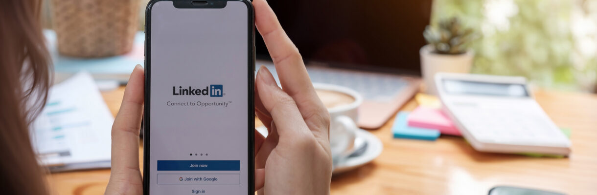 Hände halten ein Smartphone mit geöffneter LinkedIn-App vor einem Arbeitsplatz mit Laptop und Kaffeetasse.