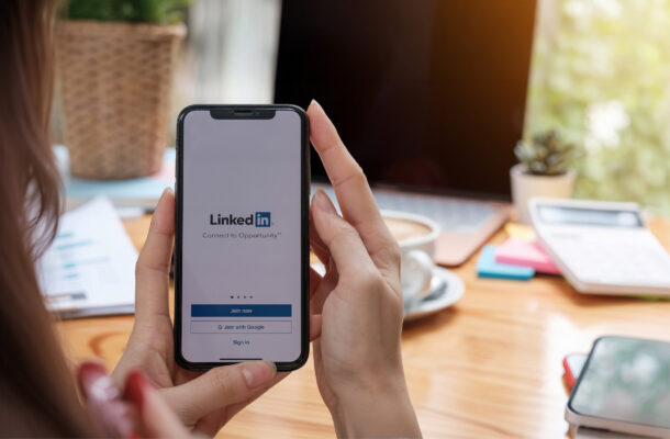 Hände halten ein Smartphone mit geöffneter LinkedIn-App vor einem Arbeitsplatz mit Laptop und Kaffeetasse.