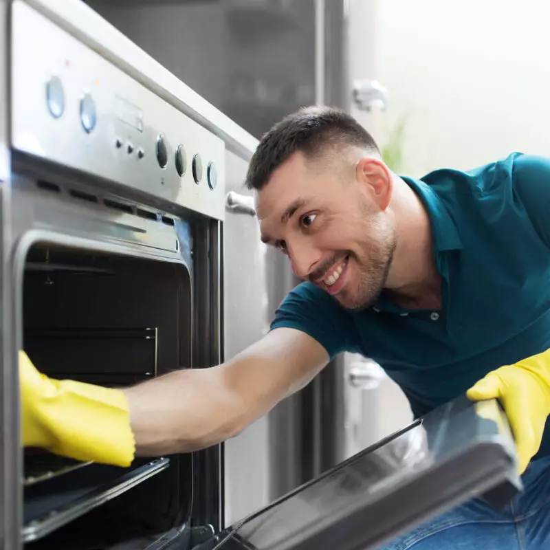 Ein Mann in einem blauen Hemd mit gelben Handschuhen putzt einen Backofen.