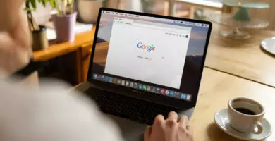 Person sitzt an einem Tisch und nutzt einen Laptop mit geöffneter Google-Startseite; daneben steht eine Tasse Kaffee in heller, wohnlicher Umgebung.