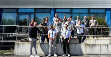 Eine Gruppe von rund 15–18 Personen mit steht bei sonnigem Wetter vor einem modernen Gebäude mit Glasfassade und lächelt in die Kamera.