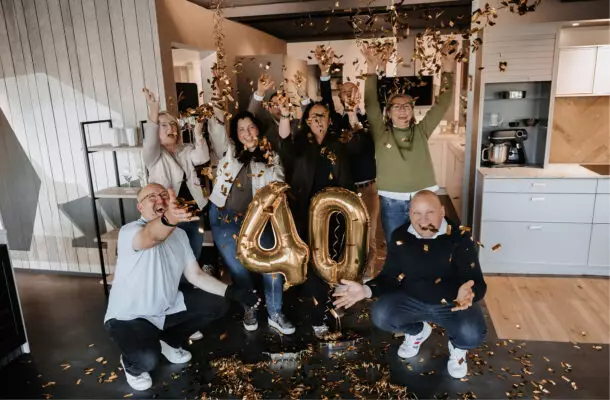 Eine Gruppe von neun Personen feiert ein 40-jähriges Jubiläum in einer modernen Küchenausstellung. Goldenes Konfetti fliegt durch die Luft, in der Mitte stehen goldene Luftballons mit der Zahl „40“, während die Menschen lachend die Arme in die Höhe strecken.