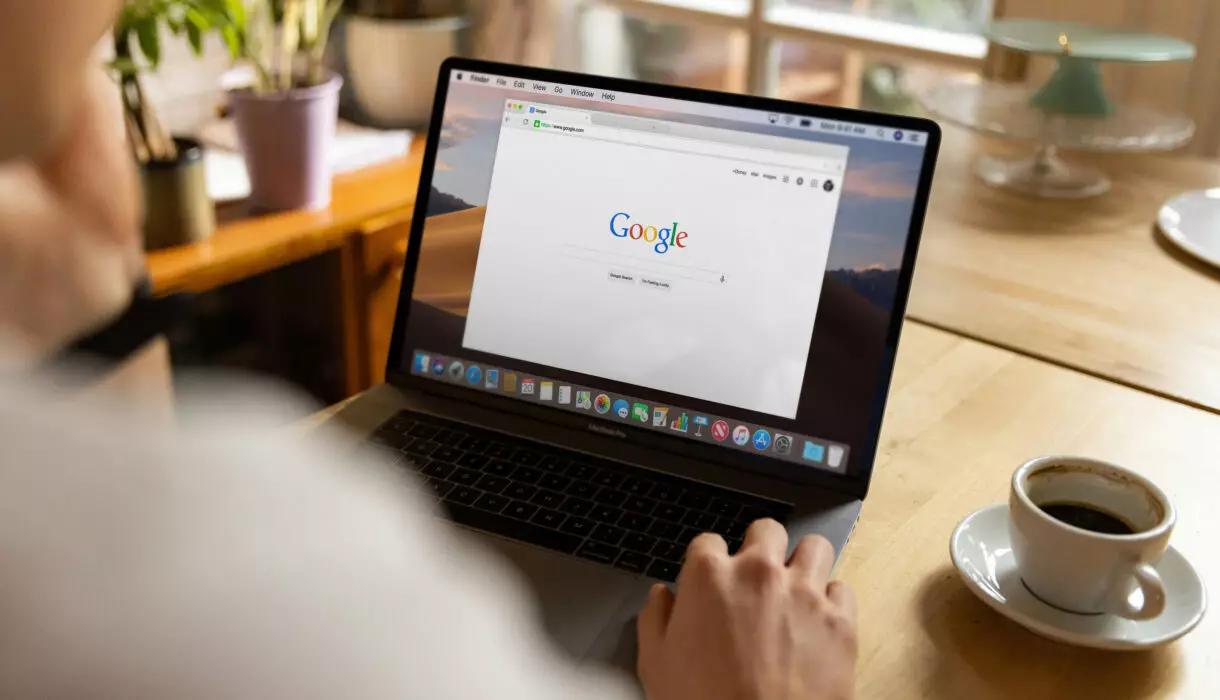 Person sitzt an einem Tisch und nutzt einen Laptop mit geöffneter Google-Startseite; daneben steht eine Tasse Kaffee in heller, wohnlicher Umgebung.