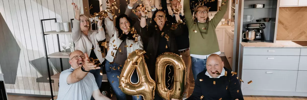 Eine Gruppe von neun Personen feiert ein 40-jähriges Jubiläum in einer modernen Küchenausstellung. Goldenes Konfetti fliegt durch die Luft, in der Mitte stehen goldene Luftballons mit der Zahl „40“, während die Menschen lachend die Arme in die Höhe strecken.