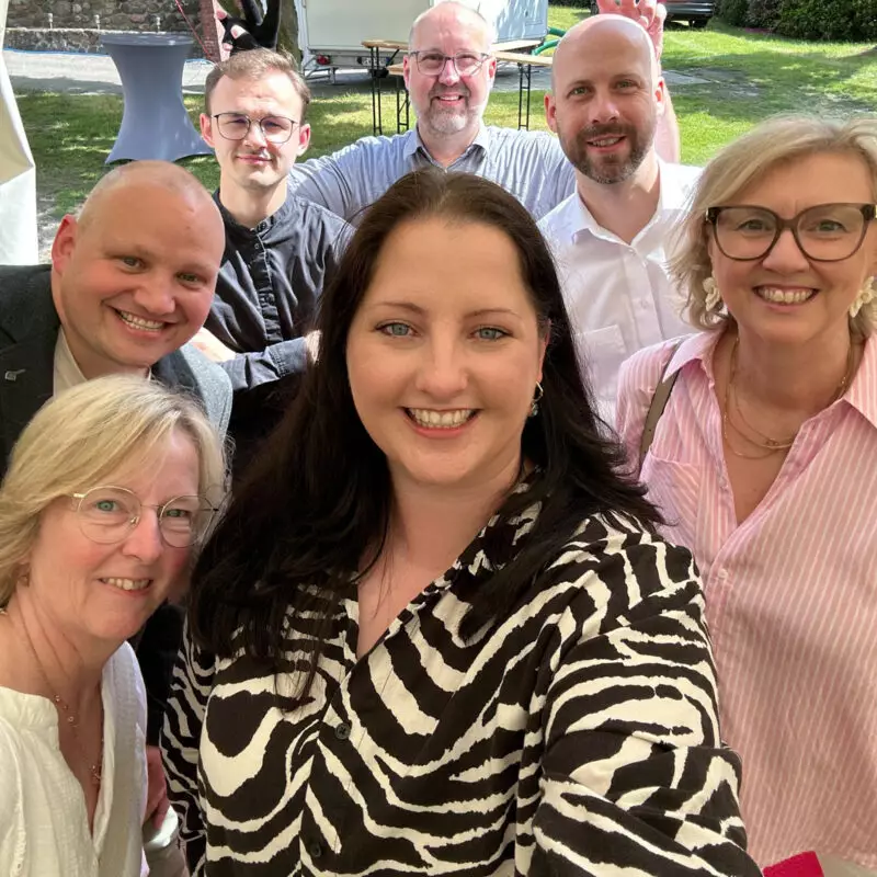 Teammitglieder lächeln gemeinsam für ein Selfie bei einer Veranstaltung im Freien. Im Hintergrund sind ein Garten, Stehtische und ein Wohnwagen zu sehen, was eine lockere und fröhliche Atmosphäre vermittelt.