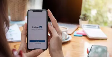 Hände halten ein Smartphone mit geöffneter LinkedIn-App vor einem Arbeitsplatz mit Laptop und Kaffeetasse.
