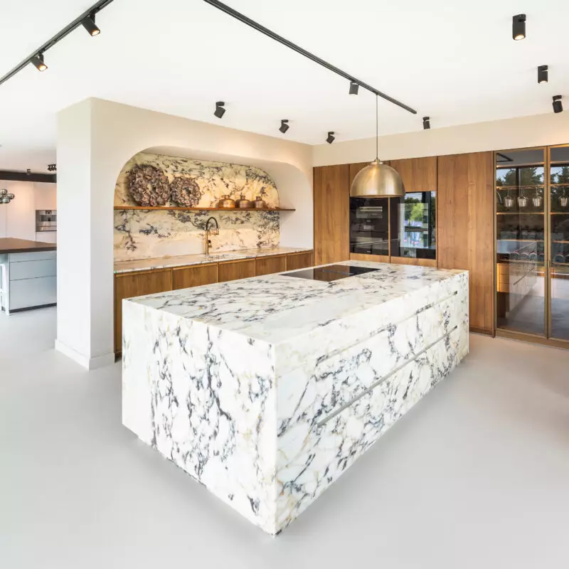 Moderne Ausstellungsküche von Mauermann Küchen mit großzügiger Kücheninsel in Marmoroptik, warmen Holzfronten, integrierten Elektrogeräten und eleganter Beleuchtung.