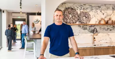Mann in blauem T-Shirt steht in einem modernen Küchenstudio von Mauermann Küchen vor einer Kücheninsel mit Marmorarbeitsplatte. Im Hintergrund tauschen sich mehrere Besucher aus.