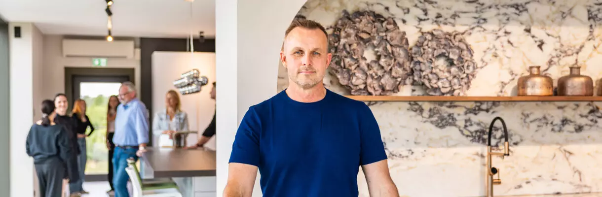 Mann in blauem T-Shirt steht in einem modernen Küchenstudio von Mauermann Küchen vor einer Kücheninsel mit Marmorarbeitsplatte. Im Hintergrund tauschen sich mehrere Besucher aus.