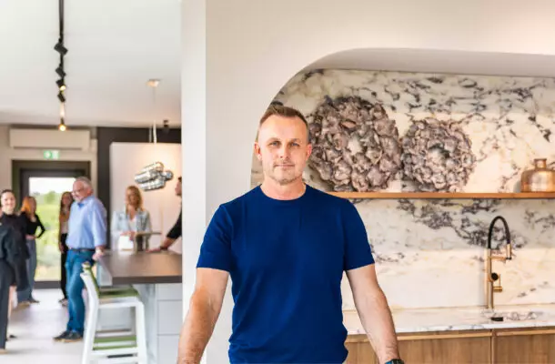 Mann in blauem T-Shirt steht in einem modernen Küchenstudio von Mauermann Küchen vor einer Kücheninsel mit Marmorarbeitsplatte. Im Hintergrund tauschen sich mehrere Besucher aus.